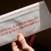 A hand holds an absentee ballot in a mail-back envelope.