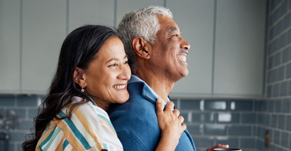 An older black couple is hugging in the kitchen.