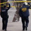 two police officers in Metropolitan Police Department jackets walk down a sidewalk with their backs to the camera holding up police caution tape as they walk under it to a crime scene