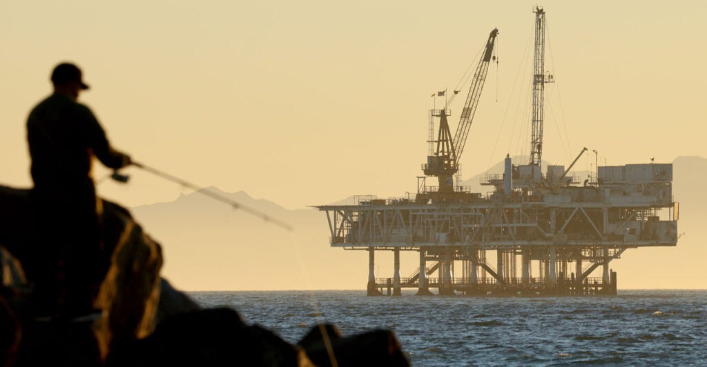 An offshore oil and gas platform in the distance with a fisherman standing on rocks and fishing in the foreground.