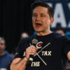 In a dark blue T-shirt with the logo “Ax the Tax,” Pierre Poilievre holds a microphone and speaks in front of a crowd during a “Spike the Hike-Axe the Tax” rally