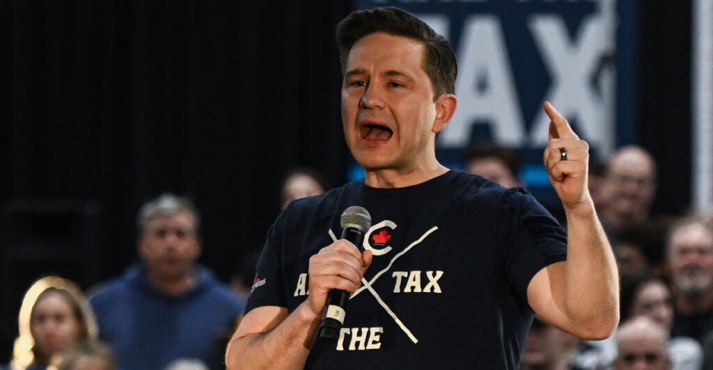 In a dark blue T-shirt with the logo “Ax the Tax,” Pierre Poilievre holds a microphone and speaks in front of a crowd during a “Spike the Hike-Axe the Tax” rally