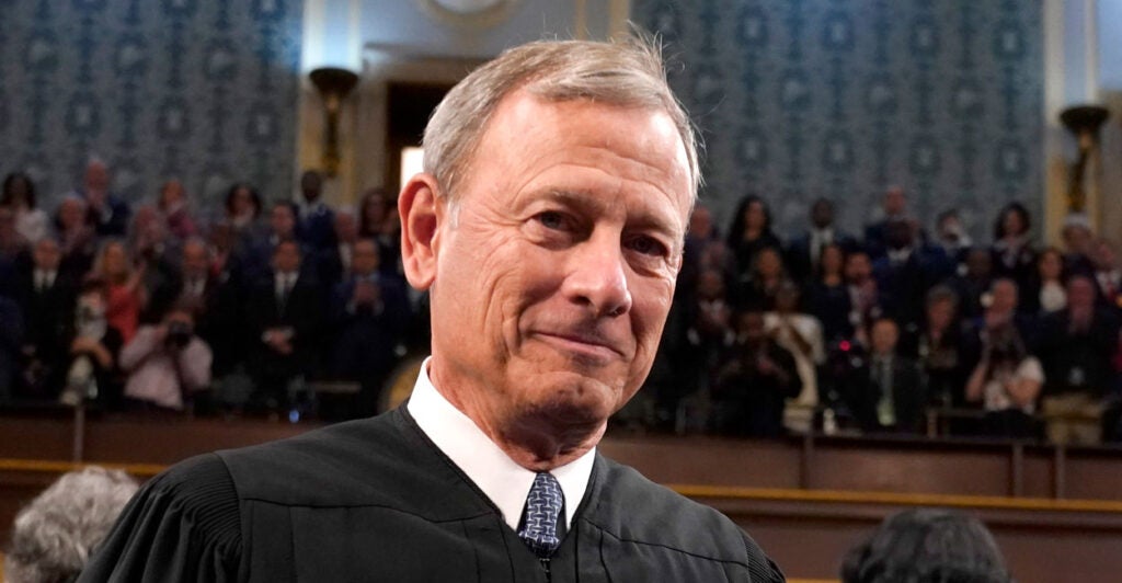 Chief Justice of the United States John Roberts Standing on the floor of the House of Representatives in a tie and black judicial robe with people filling gallery balcony behind him