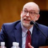 Russ Vought in a blue suit and red tie sighting at a table and speaking at his confirmation hearing