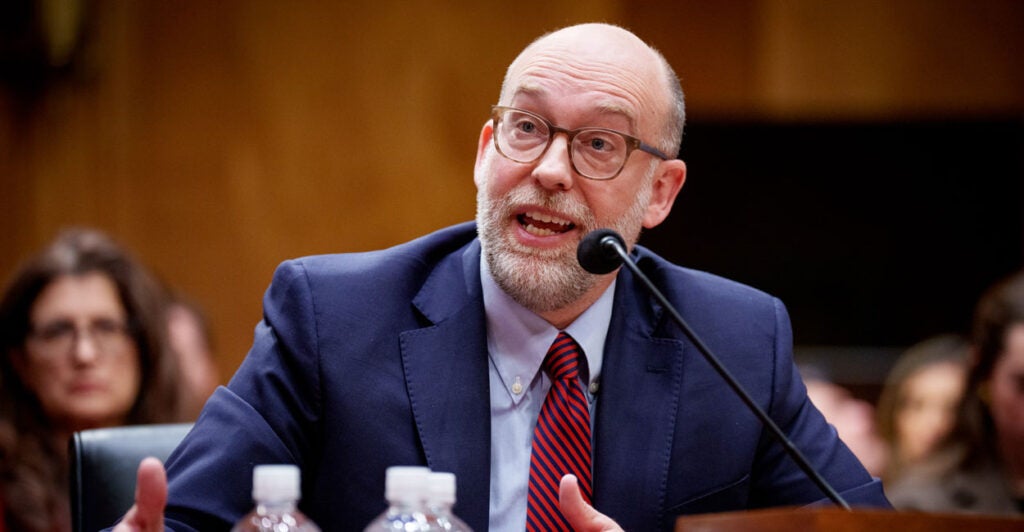 Russ Vought in a blue suit and red tie sighting at a table and speaking at his confirmation hearing