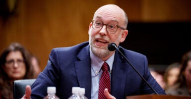 Russ Vought in a blue suit and red tie sighting at a table and speaking at his confirmation hearing