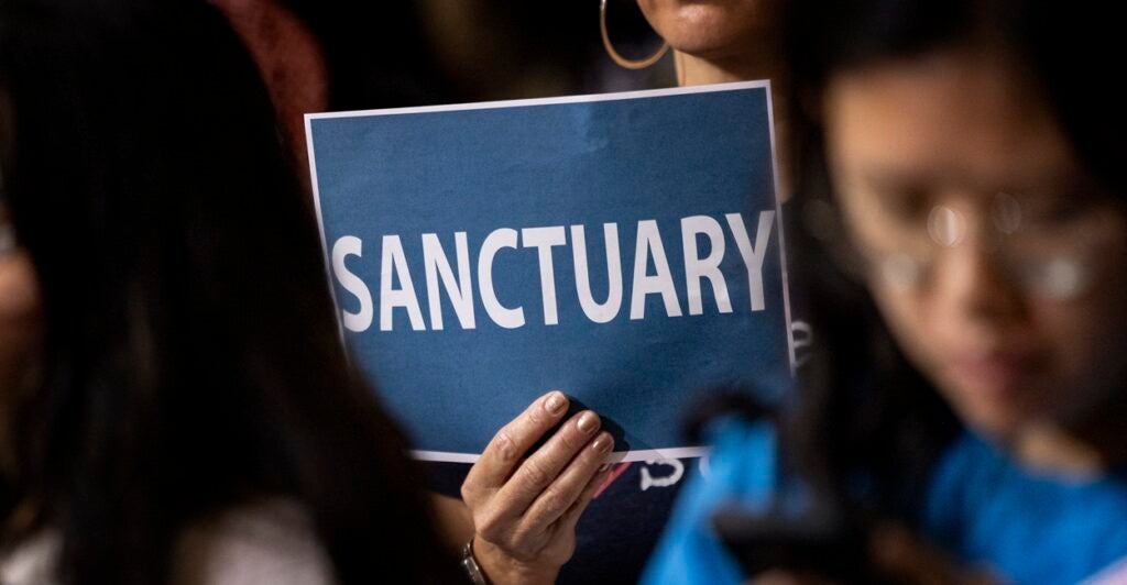 A woman holds up a sign that says "SANCTUARY" at a meeting.