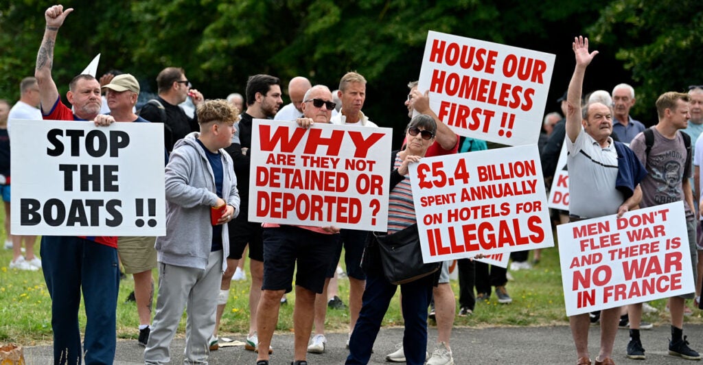 Several British men on a roadside protesting illegal aliens with large signs that say things such as "stop the boats"