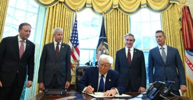 Surrounded by Cabinet members, President Donald Trump signs an executive order at the Resolute Desk in the Oval Office.