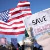 A pair of hands hold up a sign that states "SAVE USAID" as an American flag waves in the background.