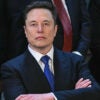 Elon Musk in a suit with a blue tie stands resolute