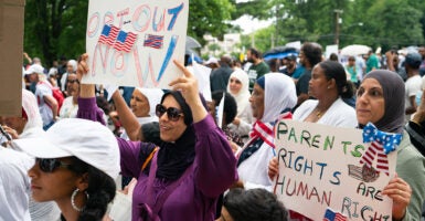 Women wearing head coverings hold signs reading "Opt-Out Now" and "Parents Rights Are Human Rights."